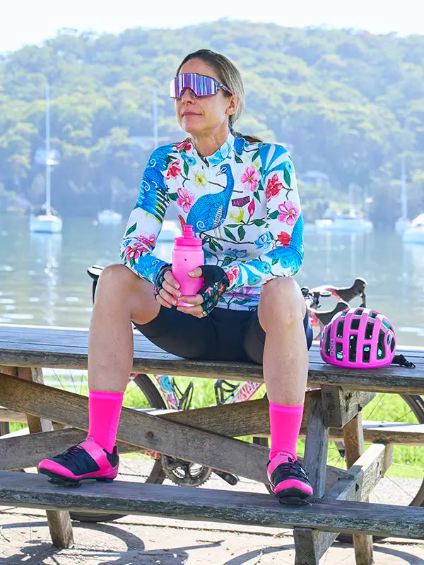 Woman sitting on a picnic table with a scenic background, wearing a Goddess White Women's Long Sleeve Summer Cycling Jersey